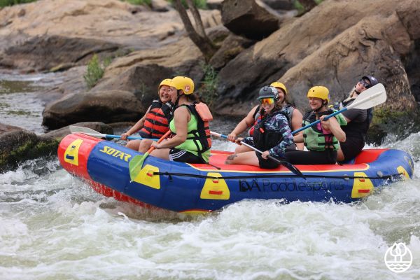 Six individuals wear helmets and life vests and hold paddles while riding in an inflatable raft. The raft is in water that is white with churning waves and flows through a rocky landscape.