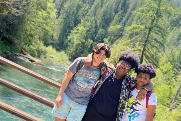 Three BIPOC boys pose together in front of a blue river surrounded by pines.