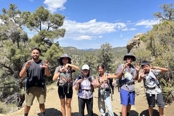 Six participants in hiking gear pose for the camera while making the numbers and letters C5LA with their hands. The scenery behind them is a desert terrain, filled with greenery despite the heat.