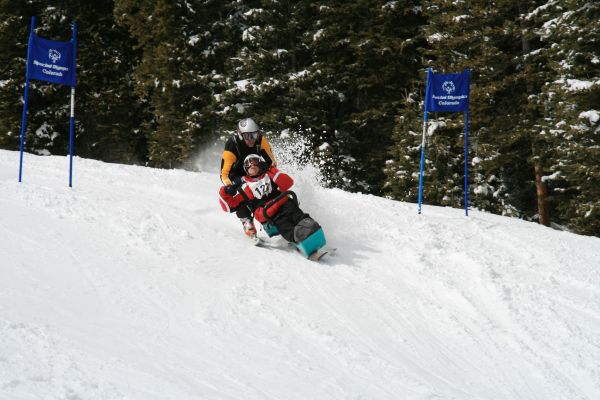 During adaptive racing, a CDA staff member guides a participant wearing a racing bib number as they slide down a snowy hill in a bi-ski.