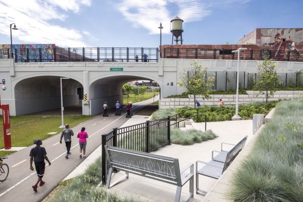 Walkers, bike riders, and segway scooter riders are sharing the non motorized roadway which is seen going under an overpass. To the right of the path are plazas with benches, cement walkways, grasses, and trees.
