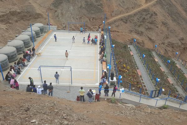 Photo shows a constructed area in a rocky hillside that features 6 cisterns, a court where people watch children playing soccer, and 3 rectangular terraced planting areas. The surrounding area appears to be arid as there is little natural vegetation.