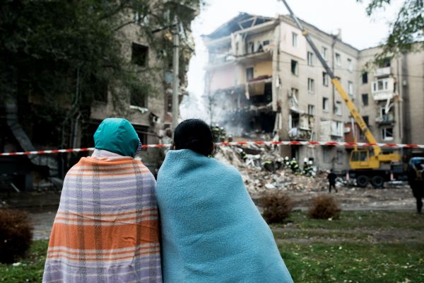 Two people with their backs to the camera have blankets draped across their backs and are looking at bombed out buildings while standing on a patch of grass.