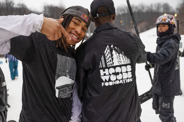 Outside in the snow, a participant faces the camera and grins as he points to the back of his peer’s jacket, which boasts the Hoods to Woods logo.
