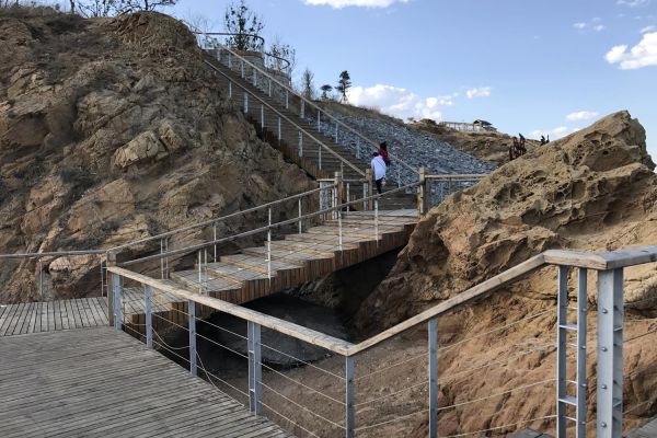 Two people walk up a wide wooden staircase. The staircase takes several turns as it goes between large rock outcrops.