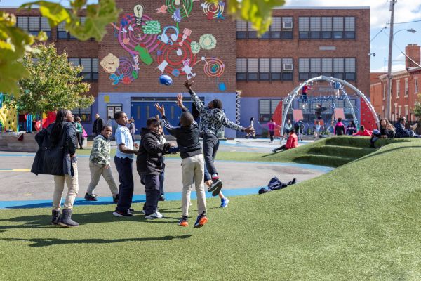A group of youth play with a football while other children sit on the berm. Behind them, kids play on the climber dome. The school building in the background is painted with bright colors and artwork.