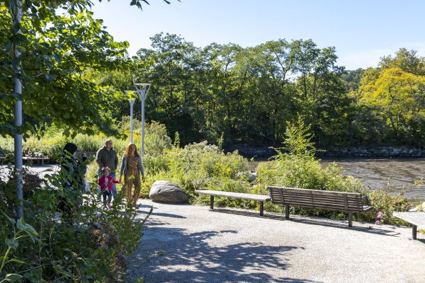 People walk along a gravel path with benches and a waterway in the background.