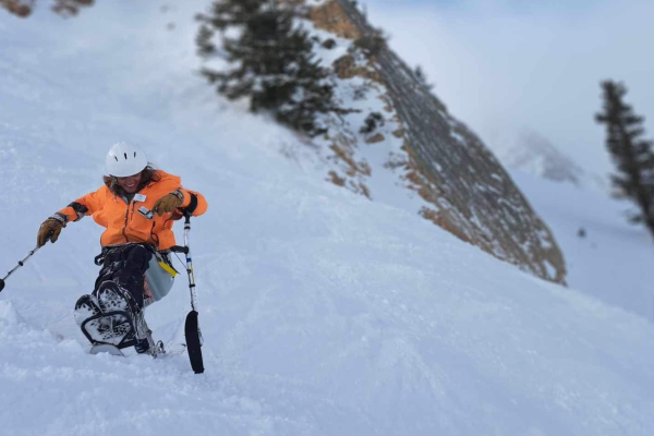 A participant in bright orange winter gear gleefully slides down a steep, snowy slope on a mono ski.