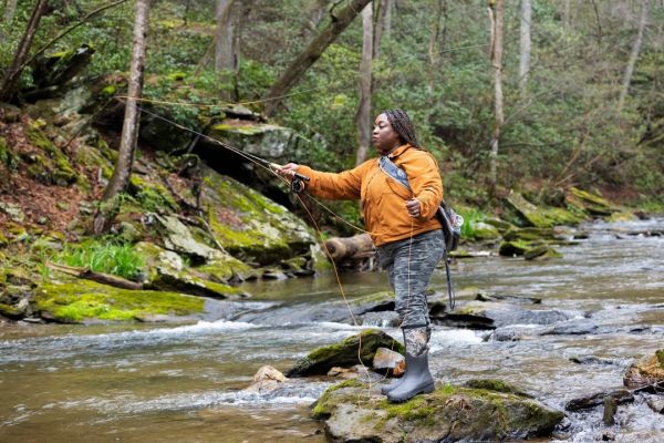 An Outdoor Afro participant stands on a mossy rock in the middle of a river and casts a fishing line. Surrounding the river bank are more mossy rocks and forest undergrowth.