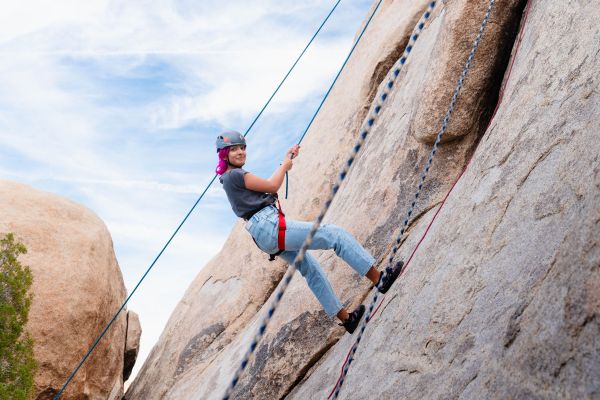 A young girl with pink hair rappels down a rock face using rope. The girl is wearing blue jeans and a helmet.