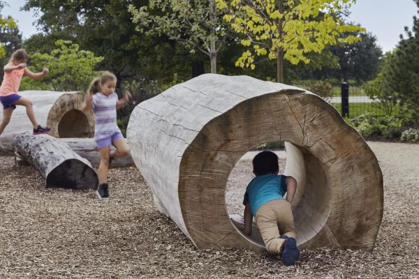 Children play in and around large, hollowed-out logs set in a flat area covered with bark mulch with trees in the background.