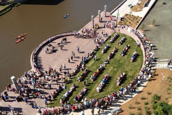 An aerial photograph shows a river on the left and a circular area consisting of half paved area and half grass with benches on the riverside. A crowd of people appear to be watching performers.