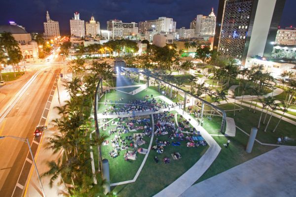 Nighttime aerial view shows the illuminated park with a crowd of people sitting on the lawn and the city skyline in the background.