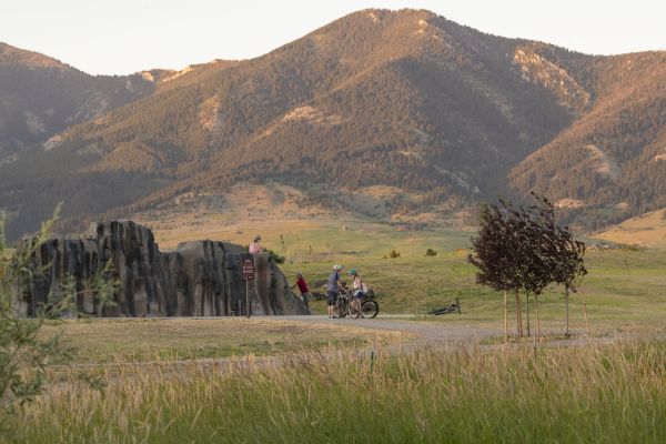 People walking and riding bicycles on a path with tall grass on either side of the path and mountains in the background.