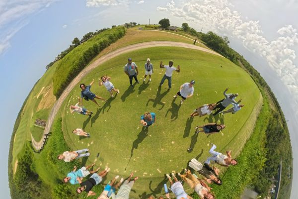 The 360 camera recreates the landscape to look like a perfect, planet-like sphere, with people standing in a grassy circle in the middle of the planet. Blue sunny sky surrounds the "planet".