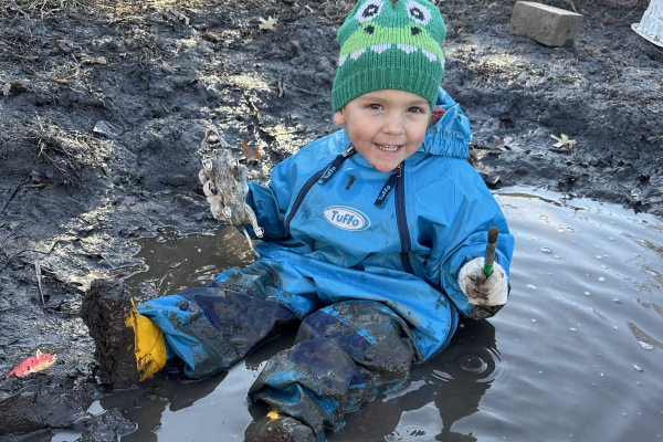 Young child in blue rain suit and green knitted cap sitting in a mud puddle smiling widely.