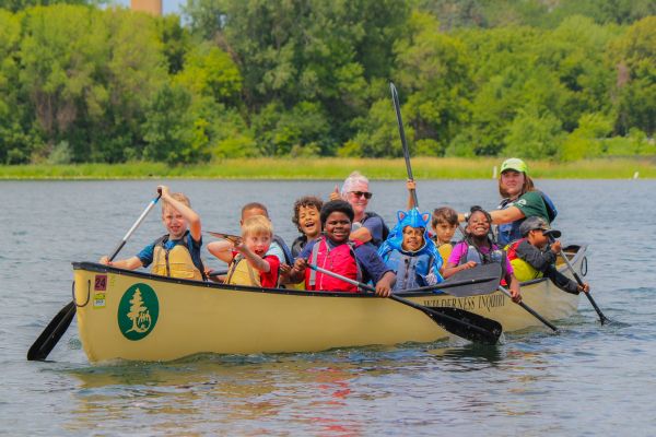 A large group of students smile while paddling their canoe during a Canoemobile program in the Twin Cities. Behind them is lush greenery that lines the edge of the lake.