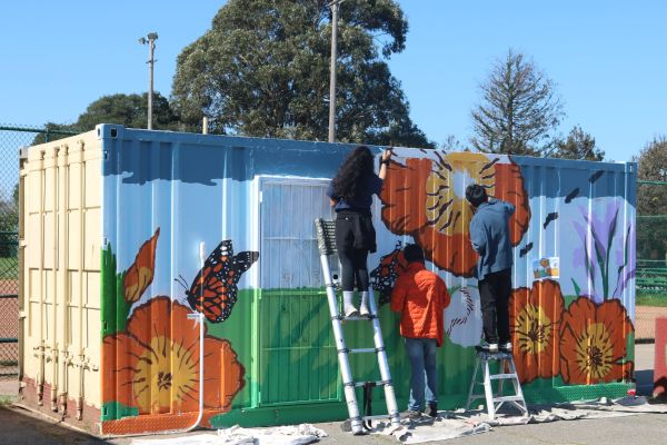 Under a bright blue sky, three youth stand on ladders painting a mural on the side of a shipping container. The mural has orange flowers and monarch butterflies amid a green and blue background.