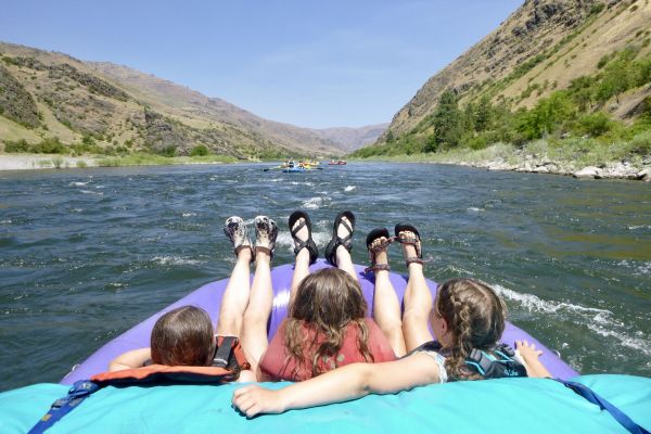Three girls lean back in their water raft and enjoy the sun as they float along a wide river. Rolling green hills line both sides of the river.