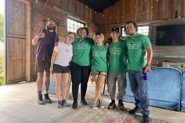 The Program Director Juleon Lewis and Farm Manager Montana Stoval line up for a picture with four Assistant Crew Leaders (ACLs) in a cabin living room. Lewis poses with a thumbs-up and a bright smile, which Stoval and the ACLs also wear on their faces.