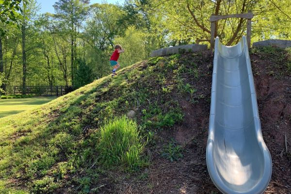 A small child runs up an earthen mound toward a hillside slide facing the camera.