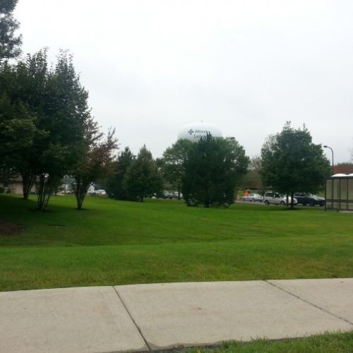 A sidewalk is in the foreground; behind is a large area of lawn, some trees, and a bus shelter. 