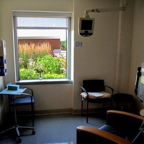 A small room with a medical screen suspended from the ceiling features a window with a view of a lush garden.