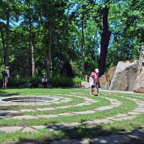 Several people walk along the spiral path in a labyrinth with trees and boulders in the background.