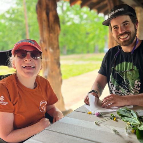 Two individuals sit at a picnic table and smile at the camera. The individual on the left is in a wheelchair and wearing an orange Beyond Boundaries t-shirt. On the table are various organic materials like flowers and leaves.