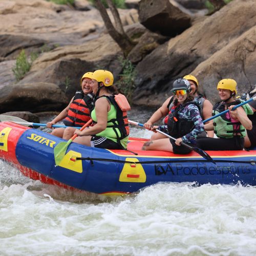 Six individuals wear helmets and life vests and hold paddles while riding in an inflatable raft. The raft is in water that is white with churning waves and flows through a rocky landscape. 