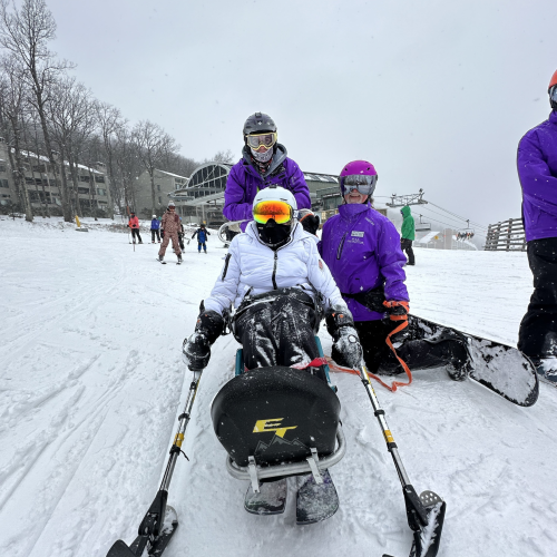 An individual in white snow gear and reflective sunglasses sits in an adaptive ski chair. Next to the individual are two people in purple snow gear, and one is kneeling and has a snowboard attached to their feet. In the background, there are other skiers atop a snowy hill. 