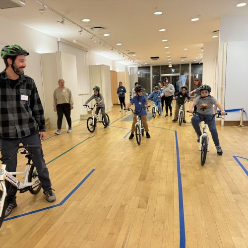 A grown man in the foreground stands with a bike in front of children on bikes in the background. Other volunteers help some children within the large, open room. All those on bikes are wearing helmets.