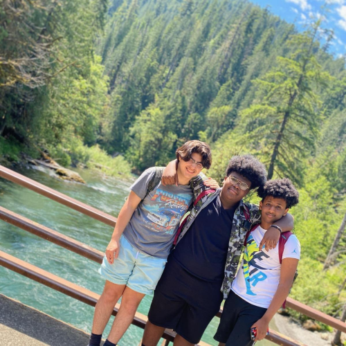Three BIPOC boys pose together in front of a blue river surrounded by pines.