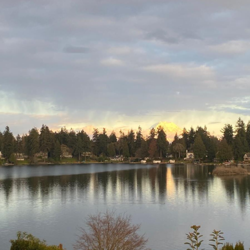 A still lake reflects the fir trees around it. The cloudy sky shows hints of orange and yellow form the obscured sun.