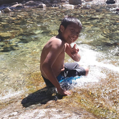 A young BIPOC boy sits in swim trunks in a stream. He smiles and has his hand in a peace sign.