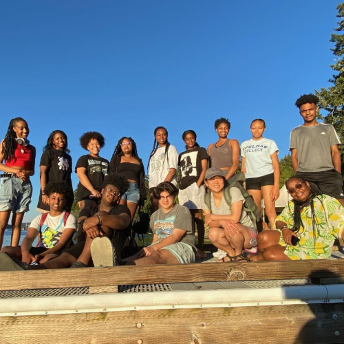 A group of BIPOC young adults pose for a photo with smiles, with some sitting on the ground and some standing behind them.