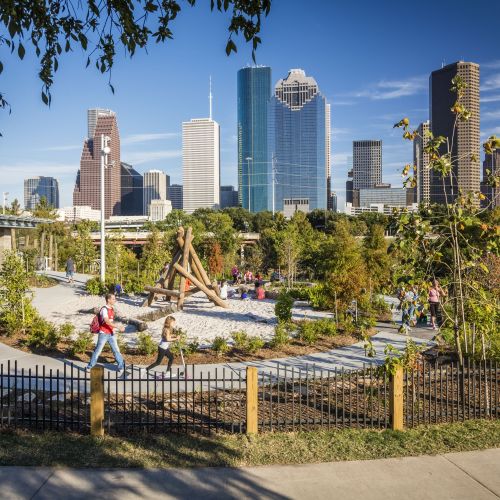 Children play in a circular, fenced-in area that features a sand-filled area, climbing structure, and paths. Tall city buildings can be seen in the background. 