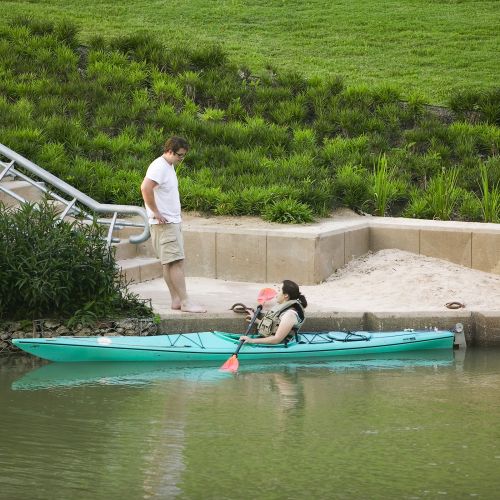 A person in a teal kayak talks with a person standing along the water’s edge with an access staircase on the left.