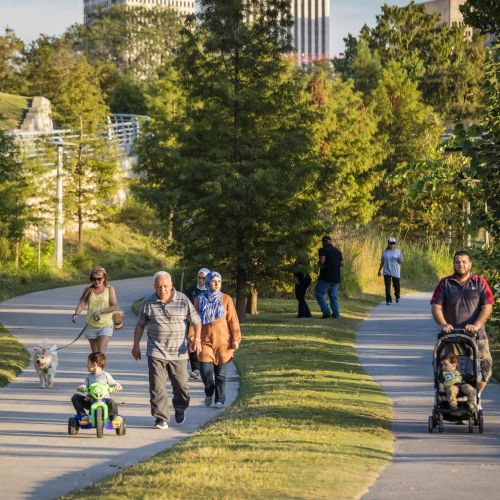 Adults, dogs, and a child on a tricycle travel along two parallel paved paths surrounded by grass and trees.