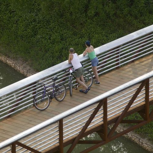 Two adults stand on the pedestrian bridge with their bicycles. The bridge overlooks the waterway and adjacent banks.