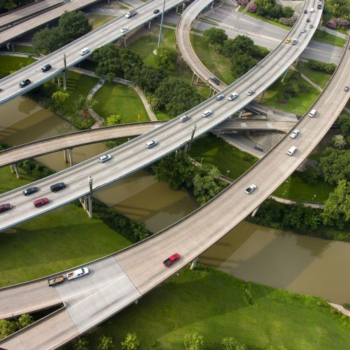 An aerial view that shows a slow-moving river flanked by grassy banks and walkways lying beneath a complex system of curving highways.
