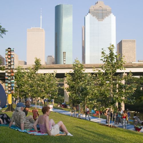 A group of people sit on the grass overlooking a path with people walking along the river’s edge. In the background is the city skyline.