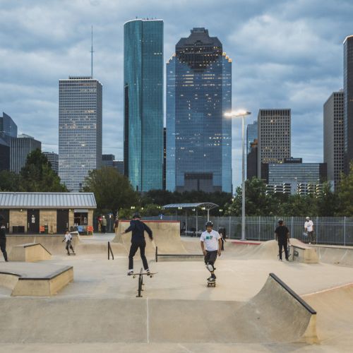 People skateboard on a concrete skateboard surface with ramps; city skyline in background.