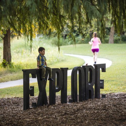 A child sits atop a sculpture that spells out the word Explore. In the background a person jogs along a path. 