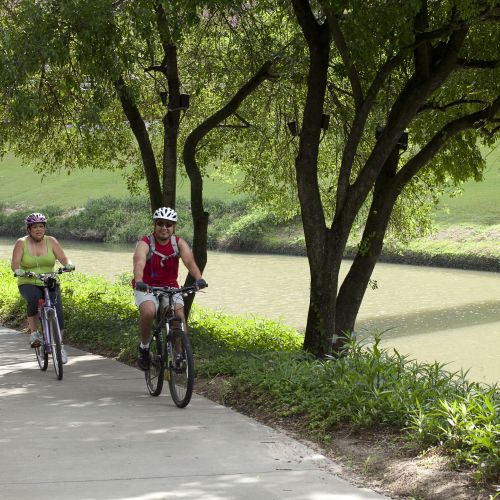 Two people on bicycles ride on a paved path that runs parallel to a slow-moving river.