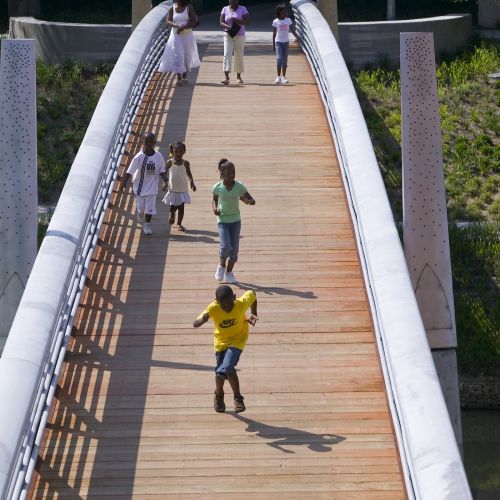Six children of various ages run and walk along a pedestrian bridge with two adults in the background. 