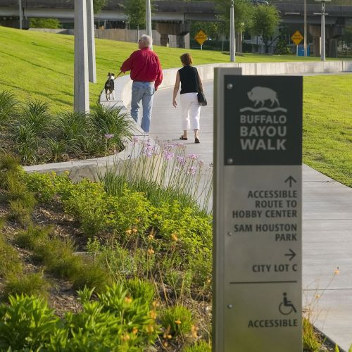 Two people and a dog walk along a paved path. A sign says it is an accessible route to the Hobby Center.