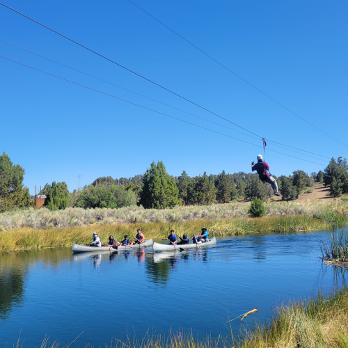 Two groups of participants paddle down in a river in separate canoes on a clear, sunny day. A person on a nearby zip line slides by with a camera.