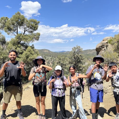 Six participants in hiking gear pose for the camera while making the numbers and letters C5LA with their hands. The scenery behind them is a desert terrain, filled with greenery despite the heat.