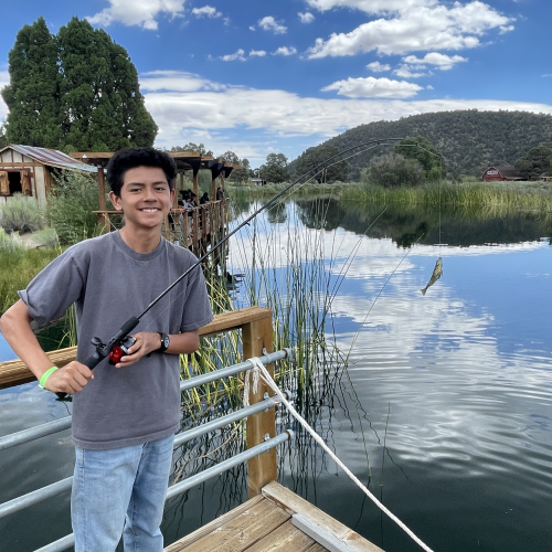 A participant wears a wide smile as he shows off the small fish he had caught, which still dangles from the end of his pole. Behind him is a clear lake that perfectly reflects the clouds in the sky.